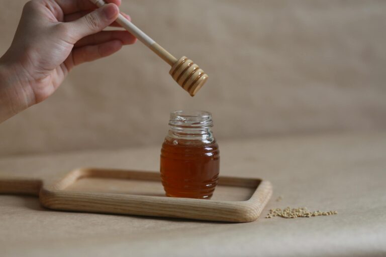 A hand holding a honey dipper over a jar on a wooden tray, showcasing the golden liquid.