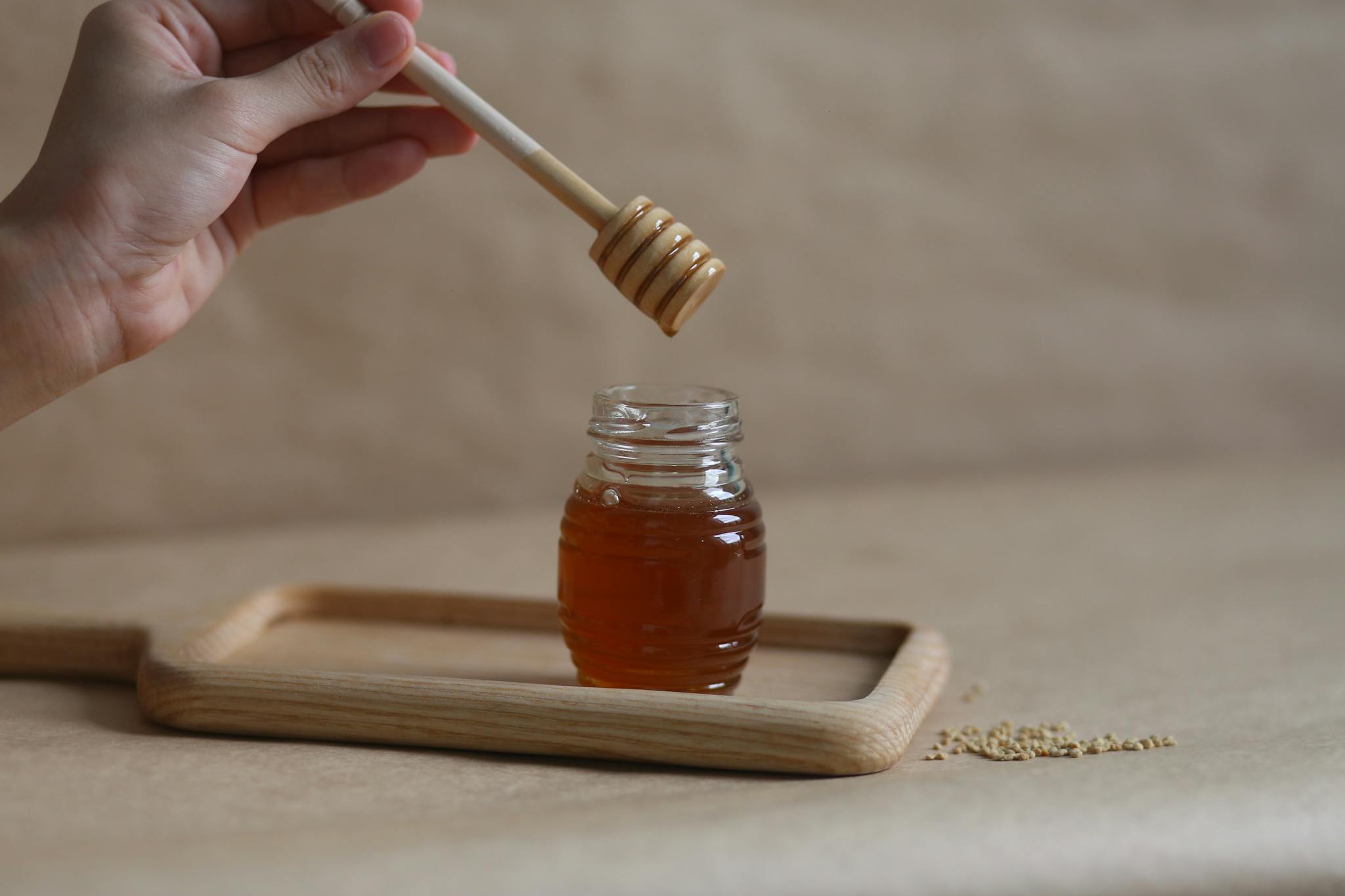 About 5 A hand holding a honey dipper over a jar on a wooden tray, showcasing the golden liquid.