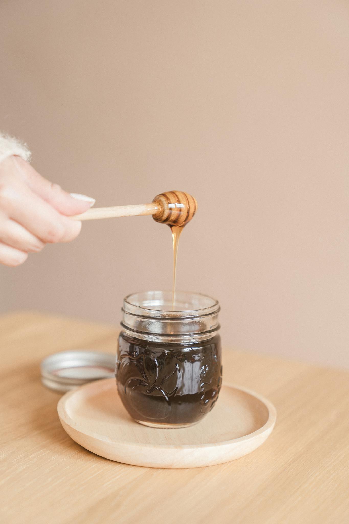 A hand holding a honey dripper over a glass jar of honey on a wooden table, creating a cozy and inviting scene.