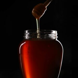 Rich amber honey dripping from a wooden dipper into a glass jar against a dark background.